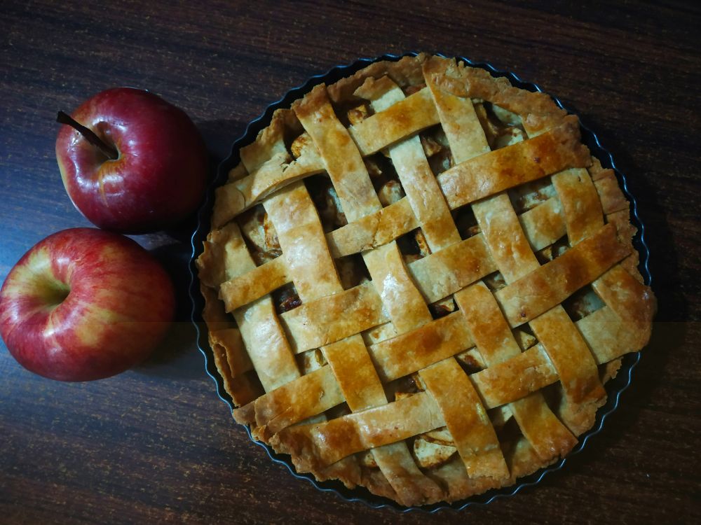 Lattice-top apple pie in a tart pan beside two red apples on a dark wooden table