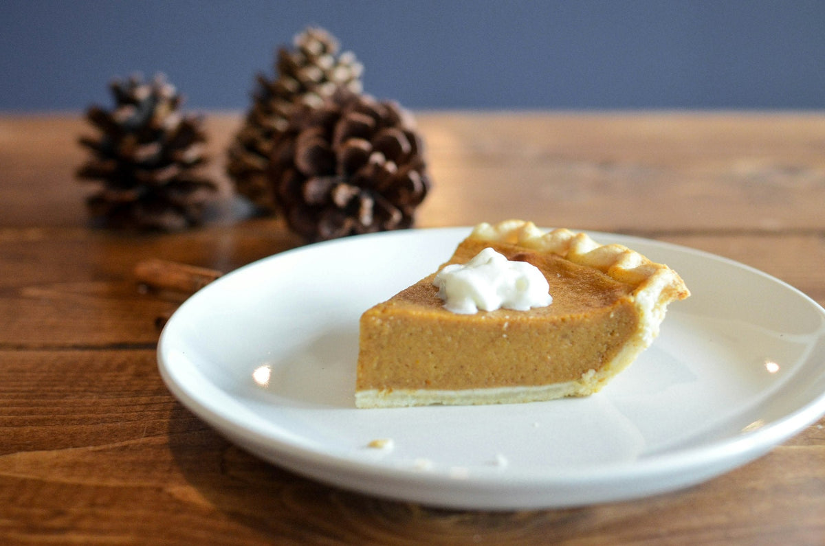 Slice of pumpkin pie topped with whipped cream on a white plate, with pinecones on a wooden table
